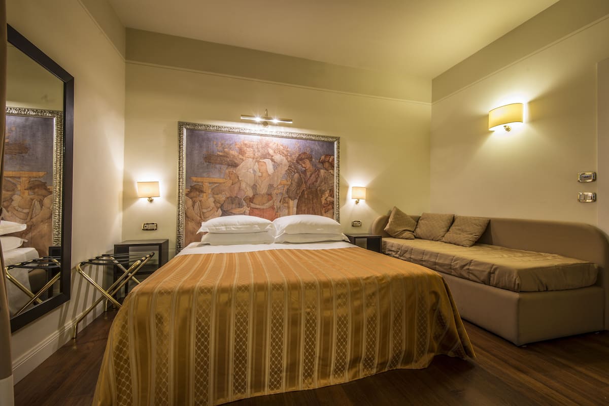 A hotel room with a large bed, a sofa, two wall-mounted lamps, and a large framed painting.