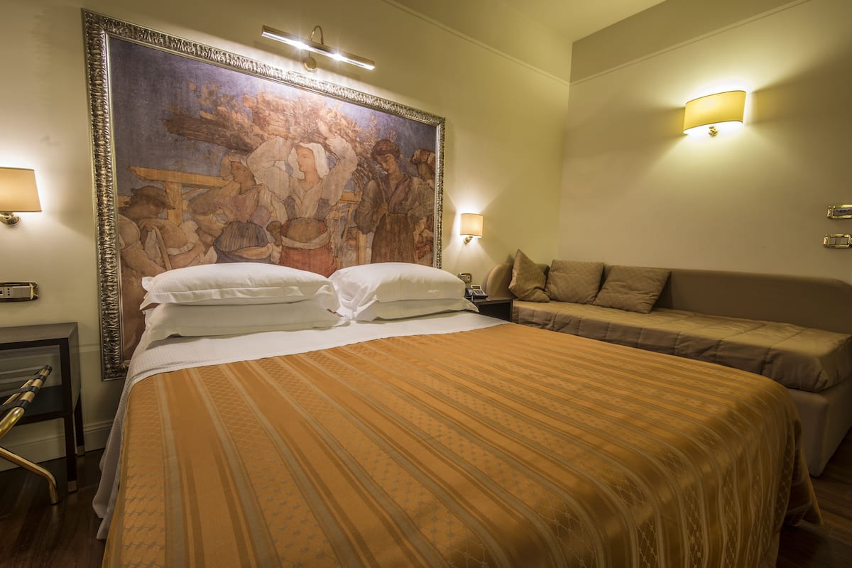 A hotel room with a large bed, a smaller sofa, a painting on the wall, and two wall-mounted lamps.