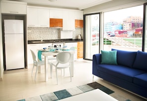 A modern kitchen with white cabinets, a black countertop, and a dining area with white chairs and a table. There is a blue sofa with teal cushions in the living area, and a large window offering a view of buildings outside. - Premium Residence Taksim Square (Istanbul)