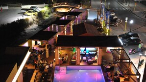 Un espacio exterior para comer con varias mesas y sillas, piscina y una zona de bar con taburetes.
