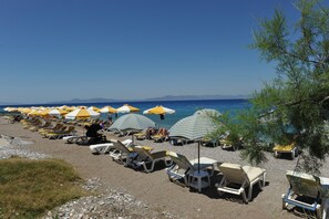A beach with rows of sun loungers and umbrellas, a clear sky, and a view of the sea and distant mountains. - Hotel Heleni Beach (Rhodes)