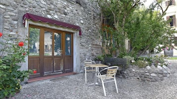 A stone building with a wooden door and windows, a gravel patio with a table and chairs, and a garden with flowers and trees.