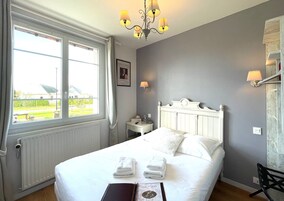 Habitación de hotel con una cama grande, una mesita de noche, una silla, una ventana con cortinas, una araña y un cuadro en la pared.