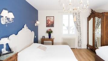 Un dormitorio con una cama blanca, una mesita de noche de madera, una pared azul decorativa, una araña, un espejo y una ventana con cortinas.