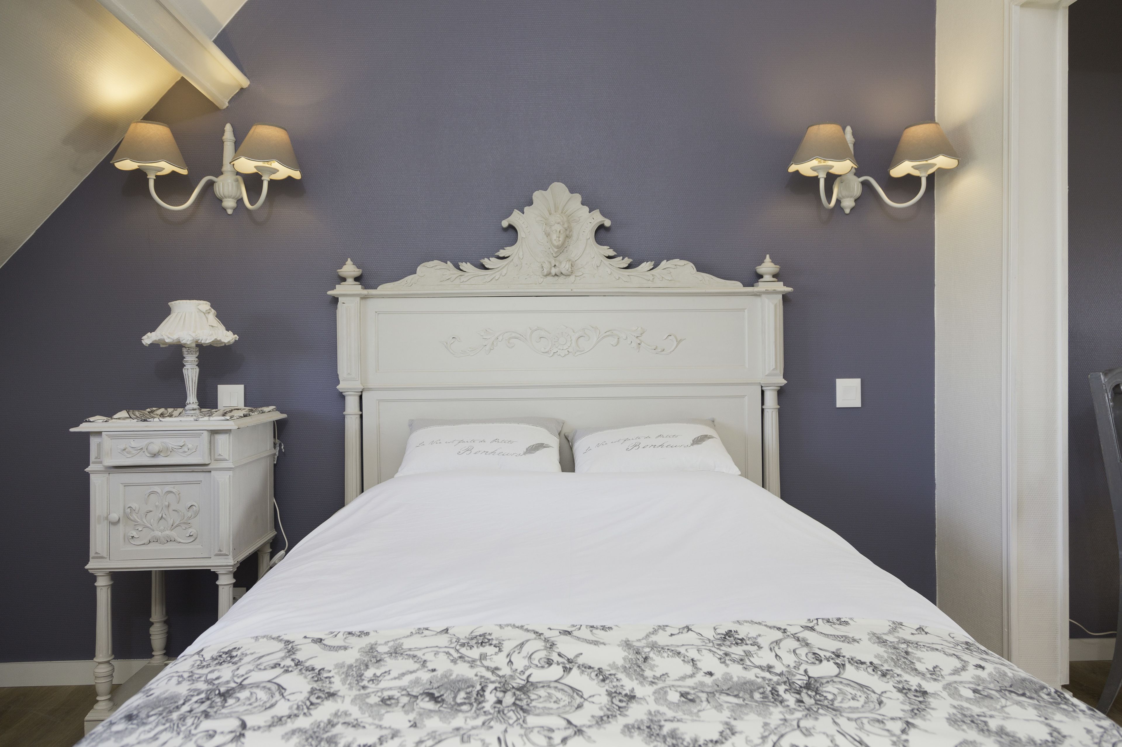 A bedroom with a white bed, two wall-mounted lamps, a nightstand with a lamp, and a decorative headboard.