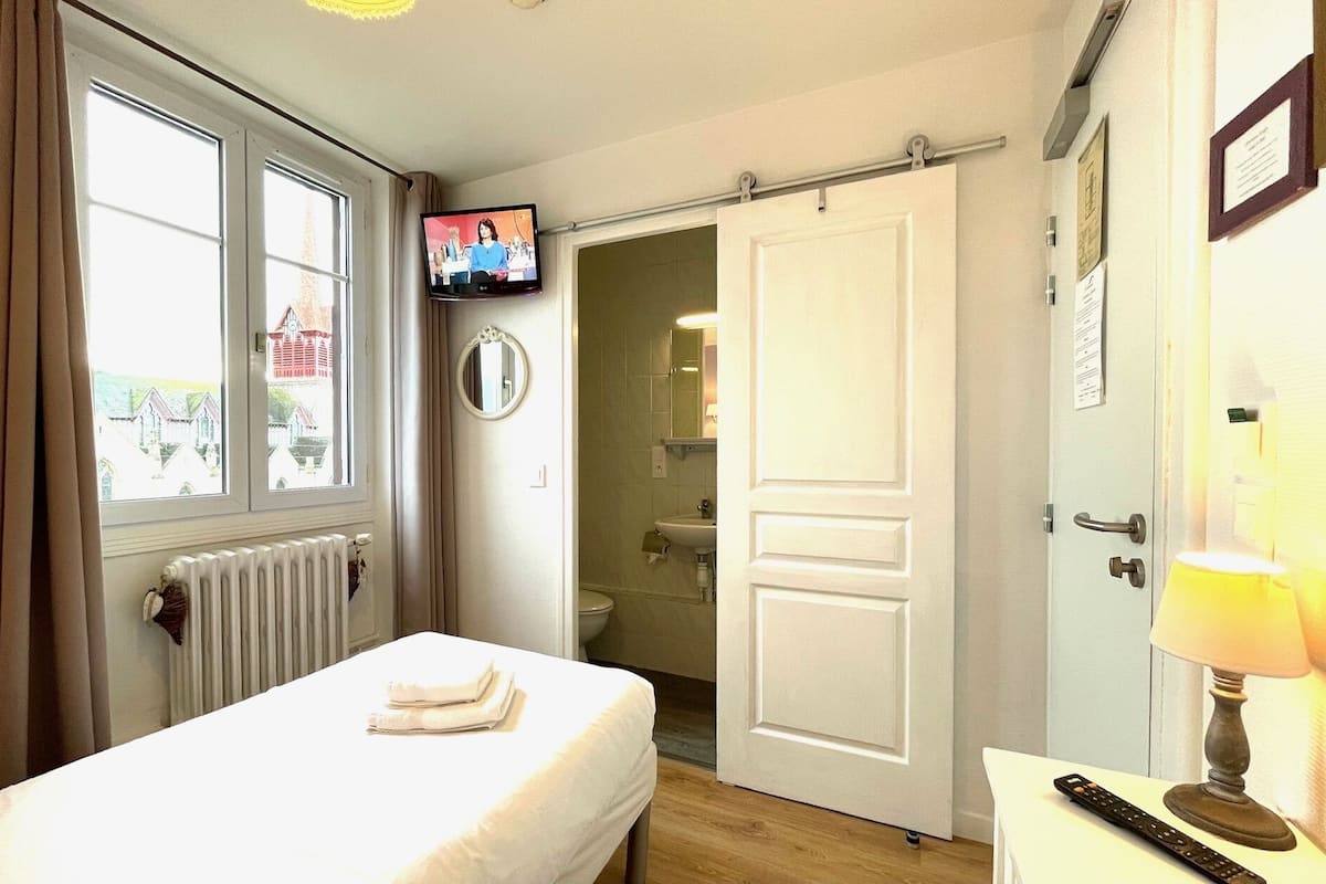 Un dormitorio con cama, televisión, puerta de baño y ventana con cortinas.