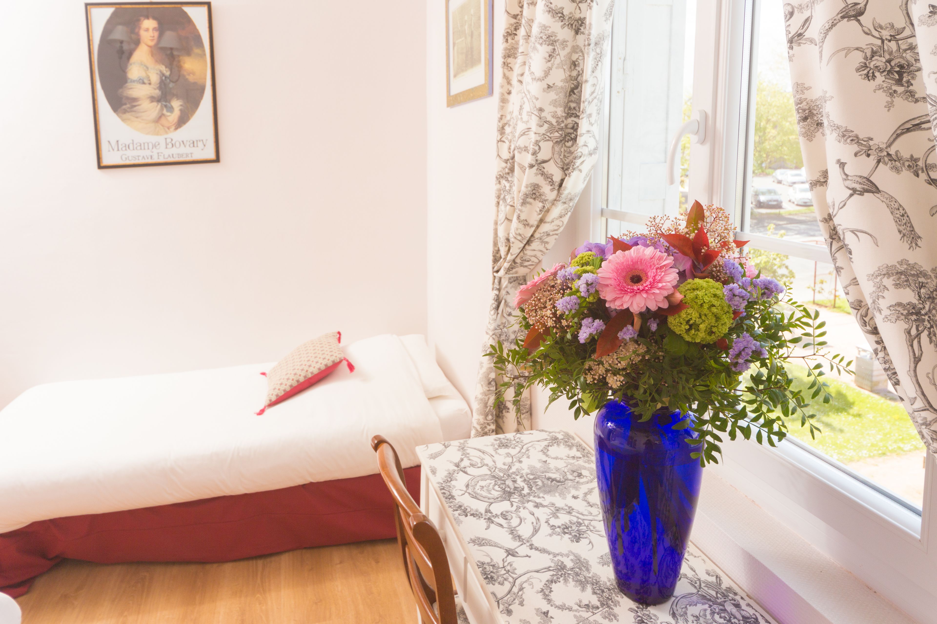 Een slaapkamer met een bed, een houten stoel, een blauwe vaas met bloemen en een raam met gordijnen.