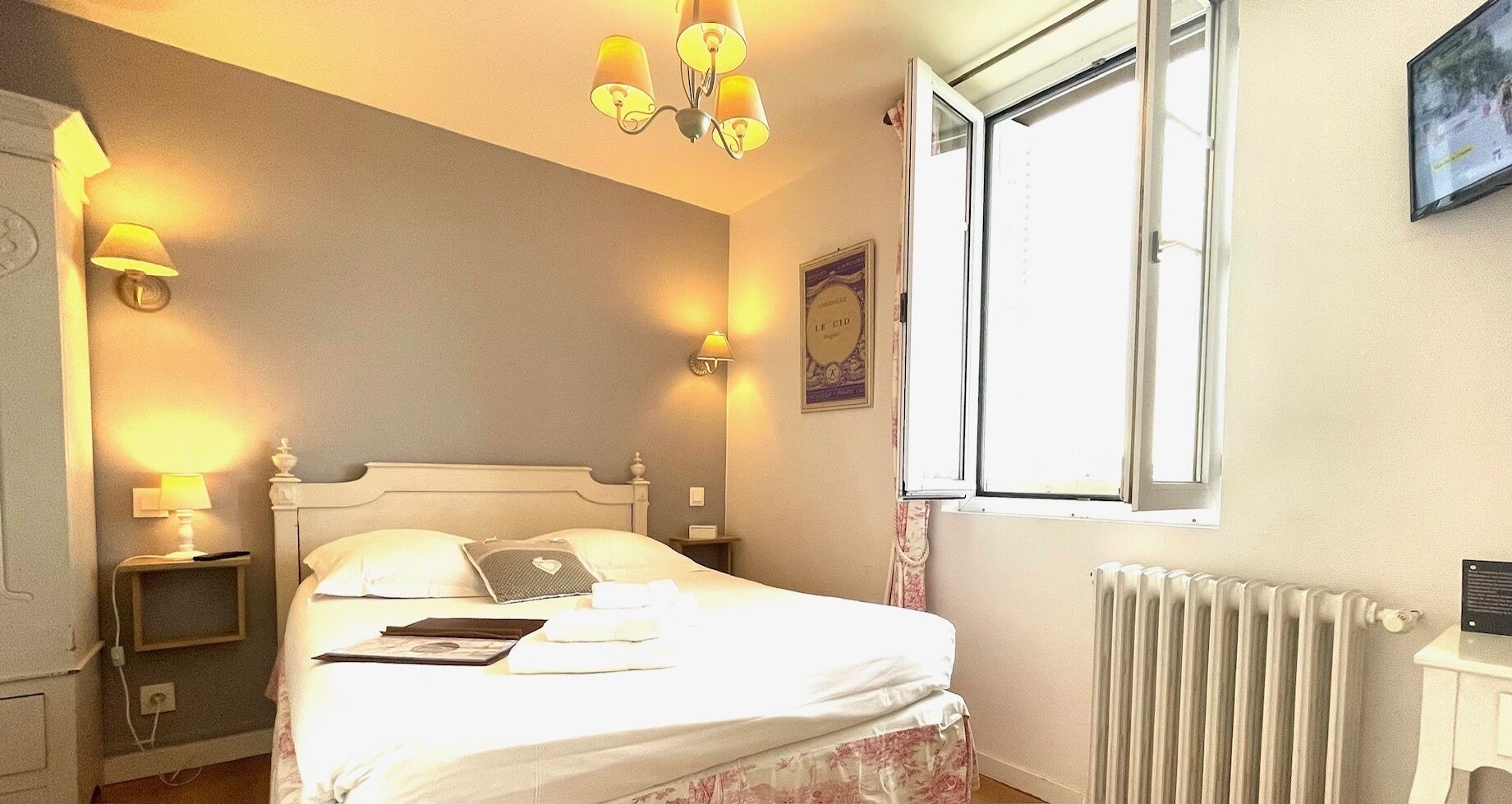 Ein ordentlich bezogenes Bett mit Kissen und einem Buch, ein Nachttisch mit Lampe, eine Heizung und ein Fenster mit Vorhängen.