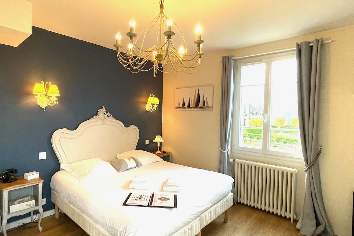 Un dormitorio con una cama blanca, un candelabro, lámparas de noche, un ventanal con cortinas y un radiador.
