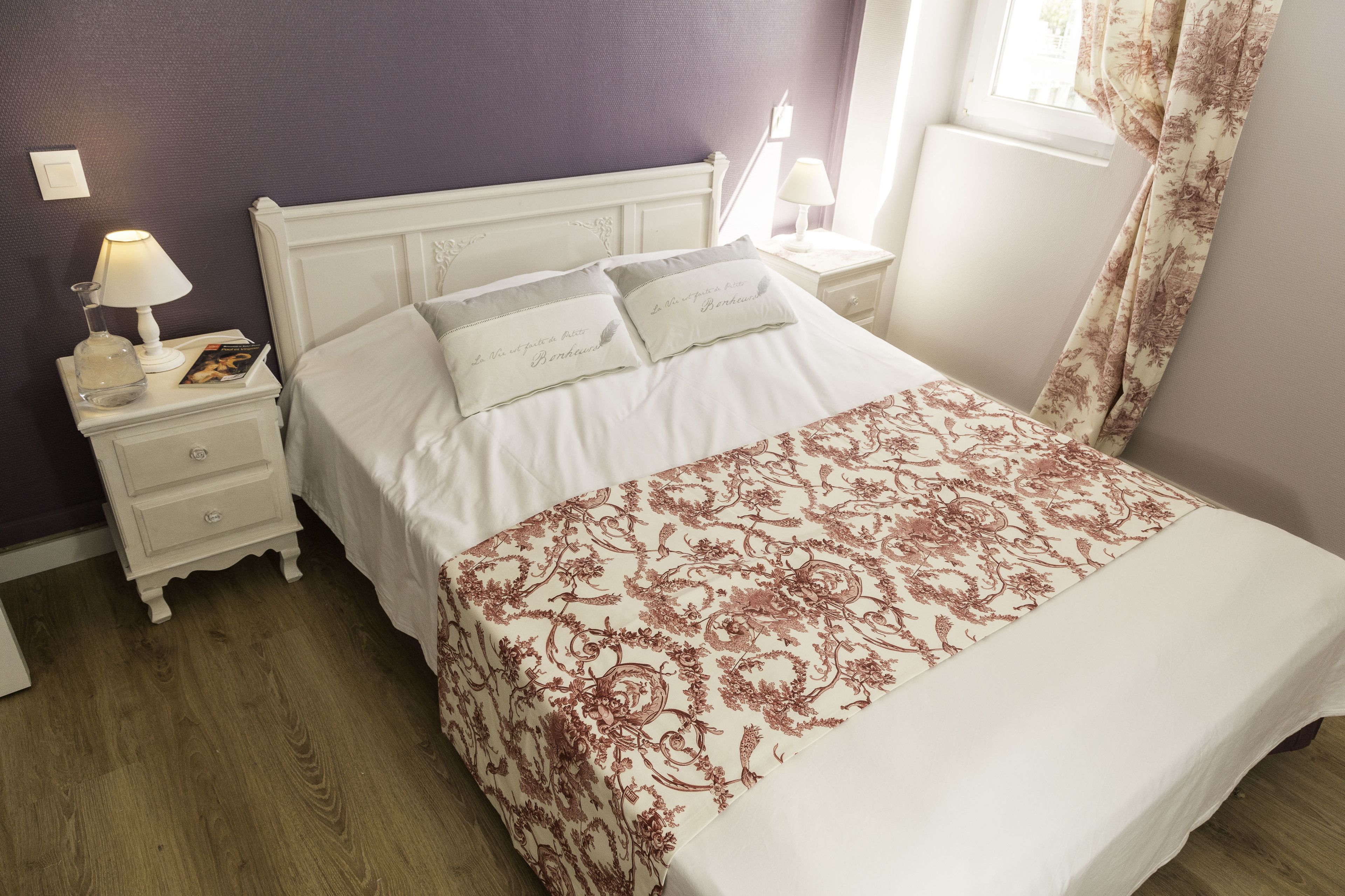A neatly made bed with a floral bedspread, a bedside table with a lamp, and a nightstand with a book and a glass.