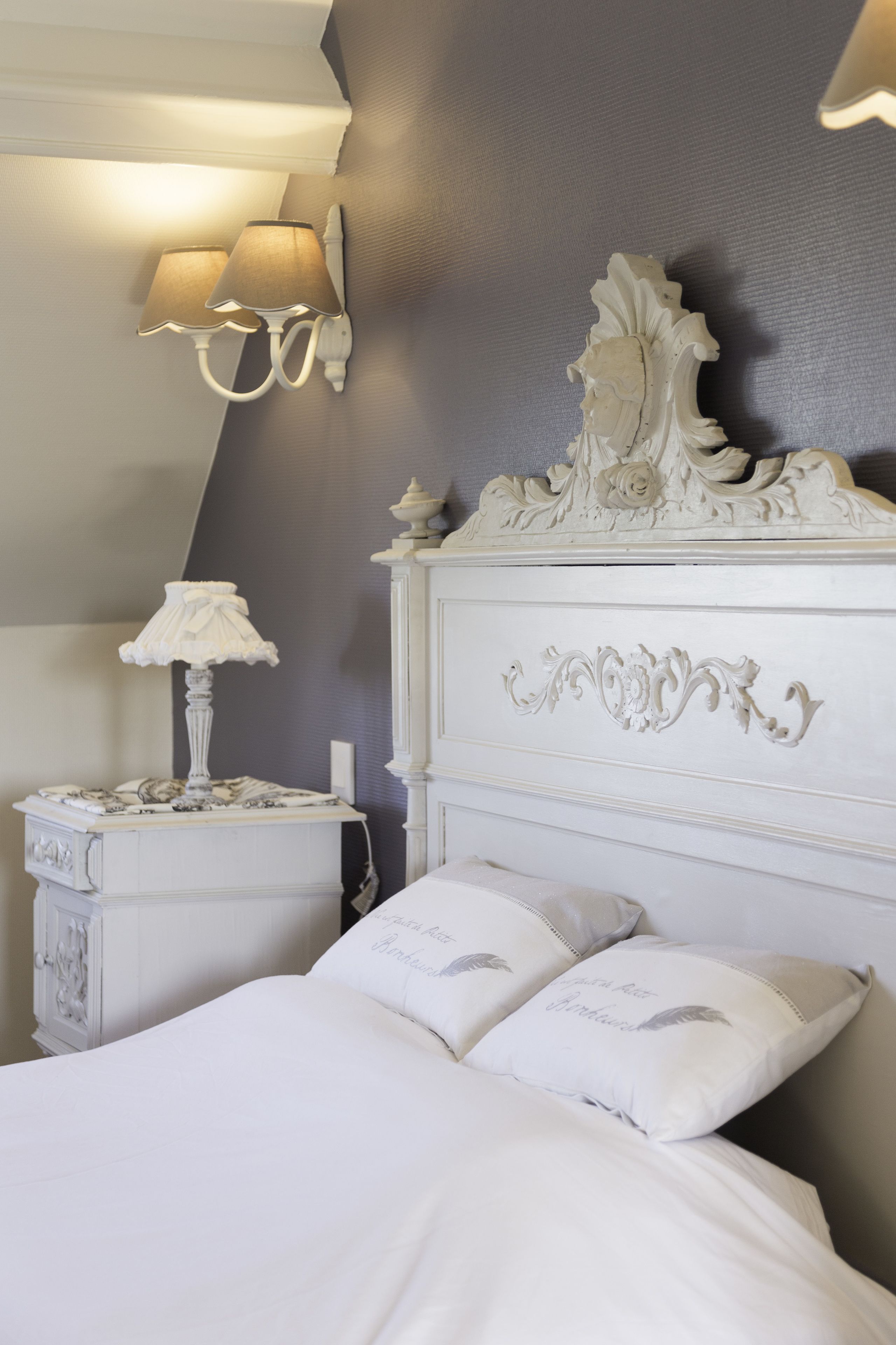 Een slaapkamer met een wit bed, een decoratief hoofdbord, wandlampen en een nachtkastje met een lamp.