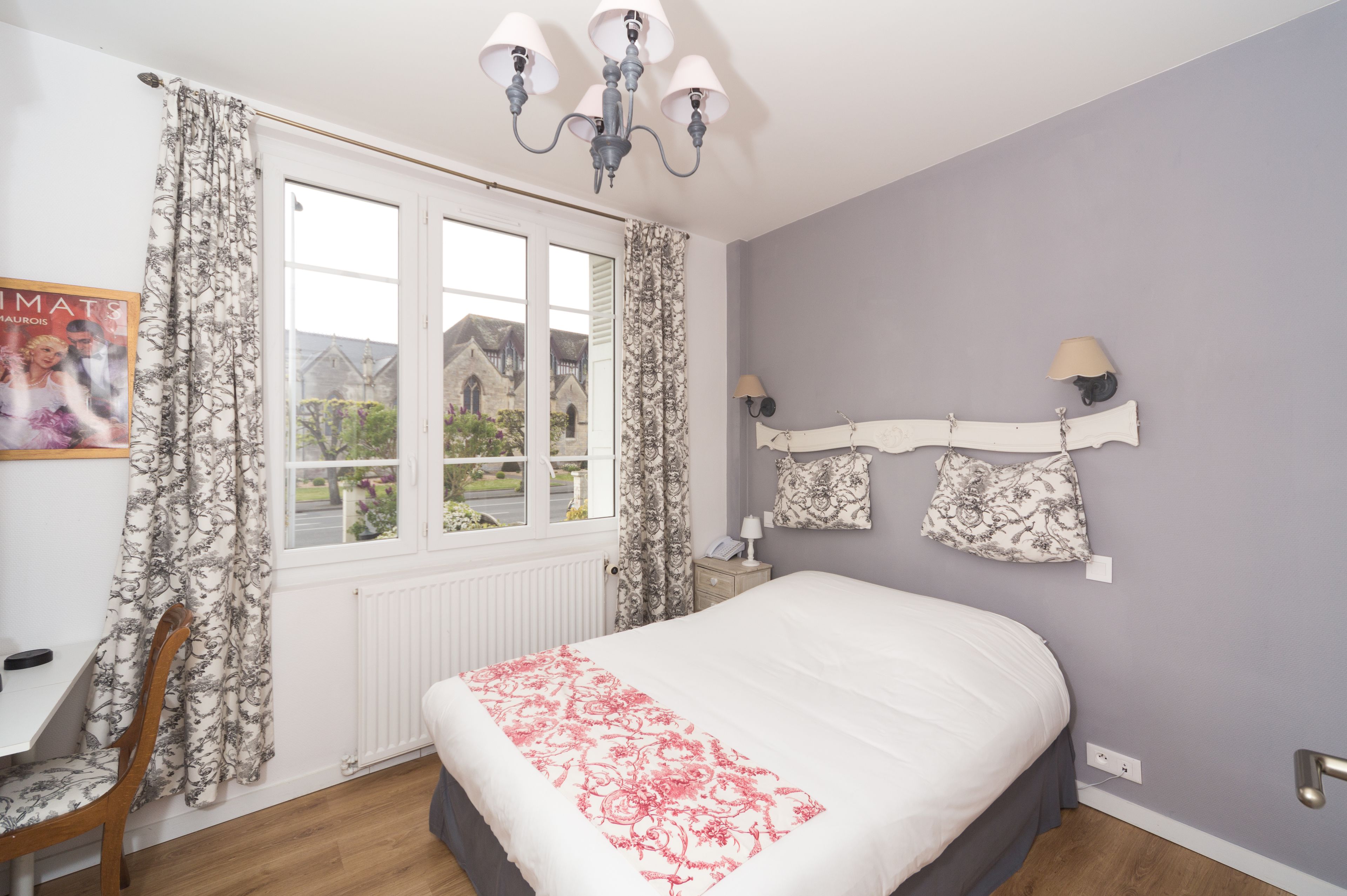 A bedroom with a large bed, a window with curtains, a chandelier, and a wall-mounted lamp.