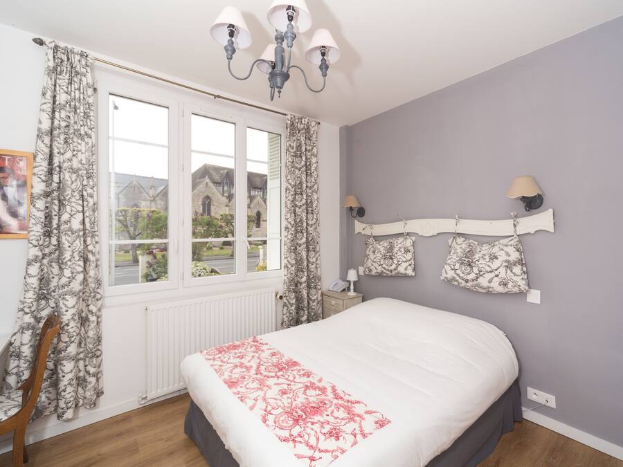 A bedroom with a large bed, a window with curtains, a chandelier, and a wall-mounted lamp.