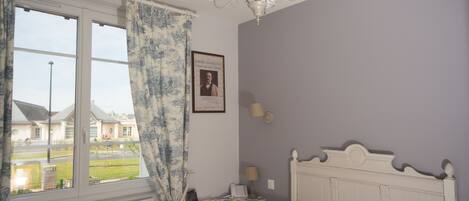 A bedroom with a bed, a bedside table, a lamp, a framed picture on the wall, and a window with curtains.