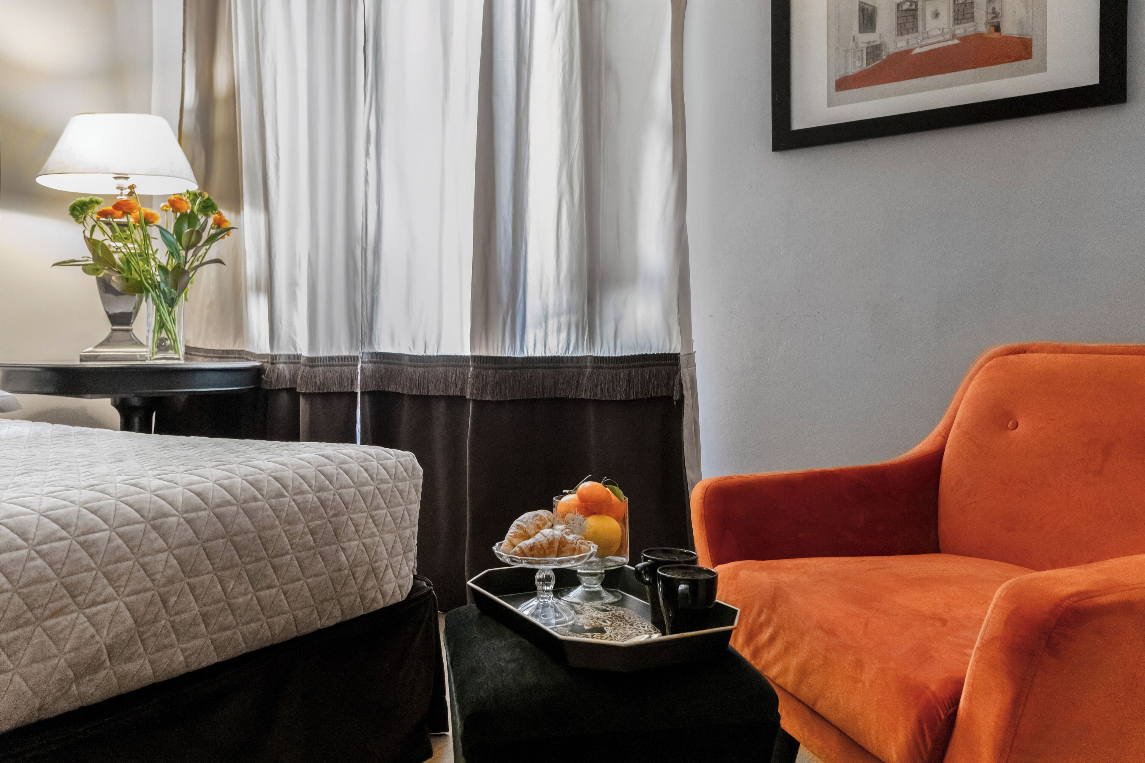 Une chambre d’hôtel avec un lit, une chaise, un plateau avec des pâtisseries et une boisson, et une lampe décorée de fleurs.