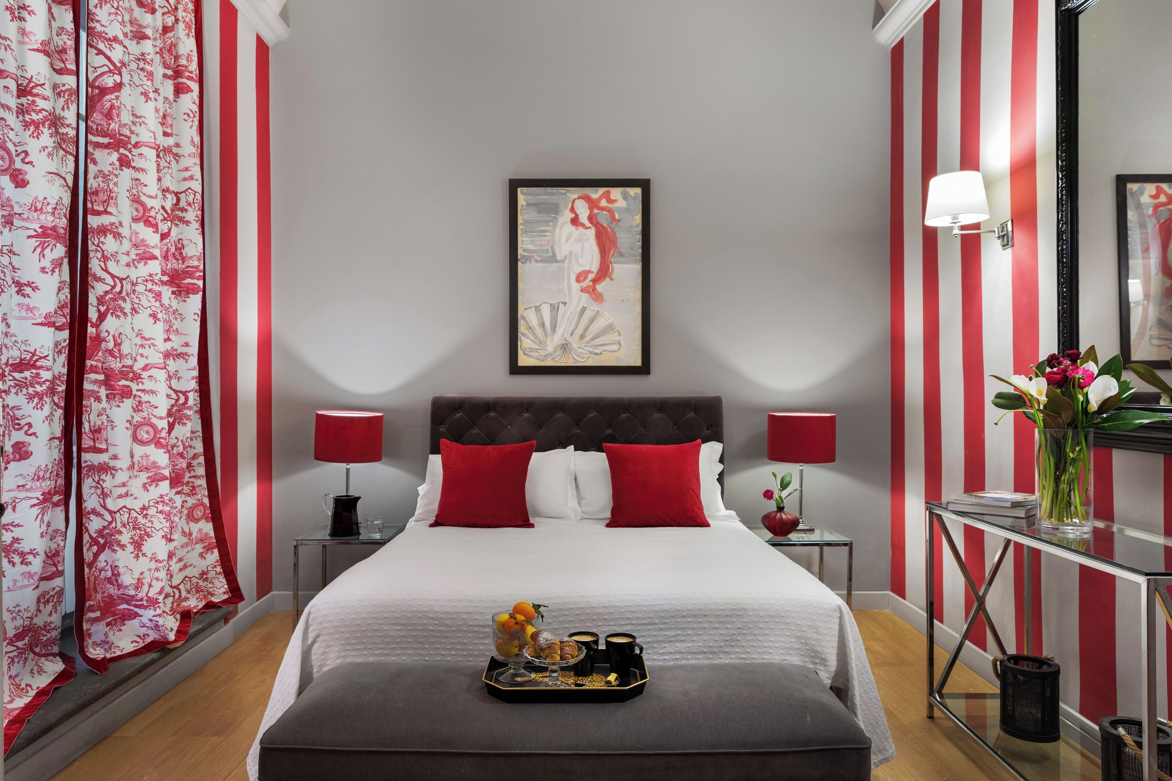A hotel room with a bed, two red pillows, a framed artwork above the bed, a bedside table with a lamp, a tray with breakfast items, and a vase with flowers on a side table.