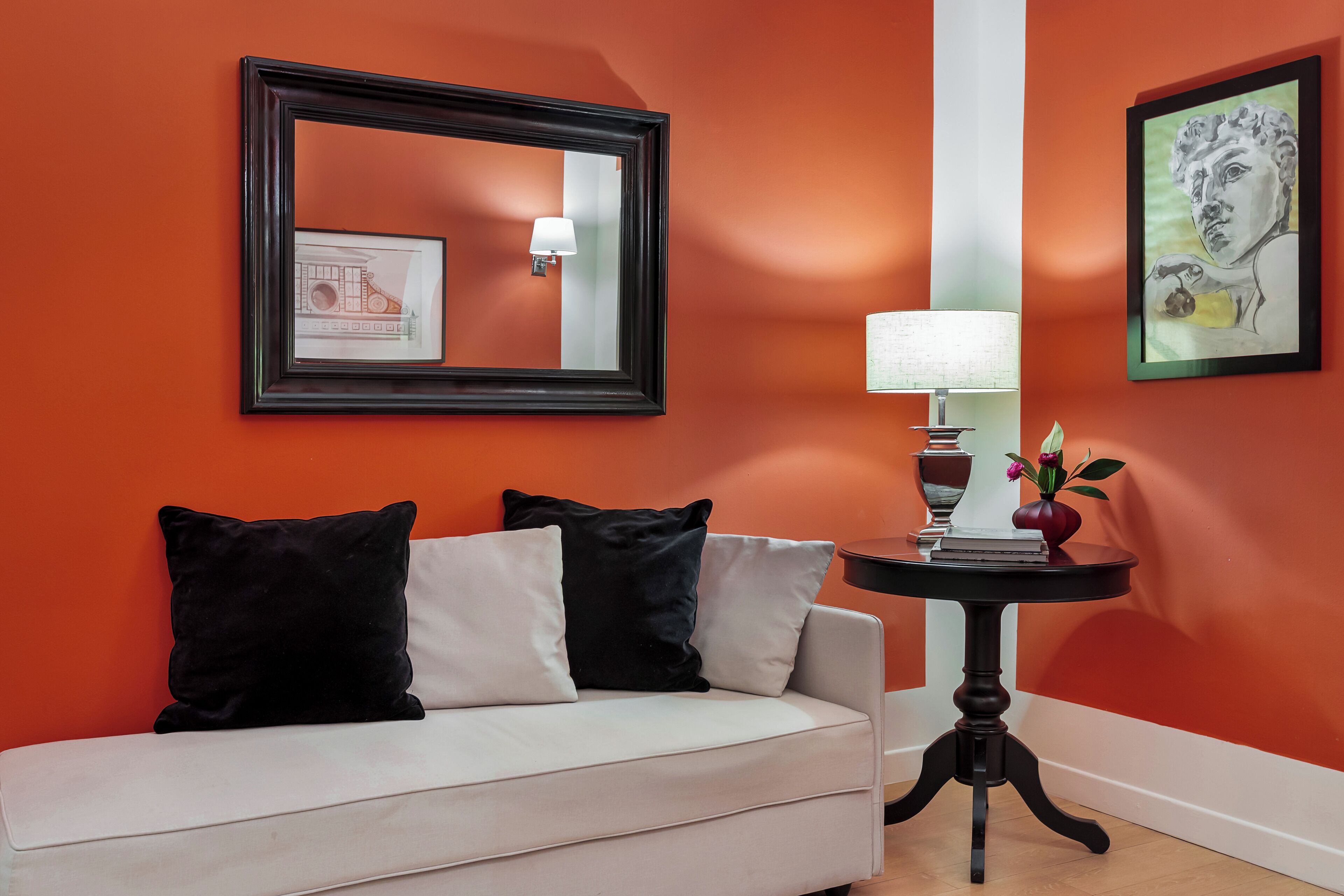 Un canapé blanc avec des coussins noirs et blancs, adossé à un mur orange. Au-dessus, on aperçoit un tableau encadré, et devant, une petite table sur laquelle sont posés une lampe et un vase.