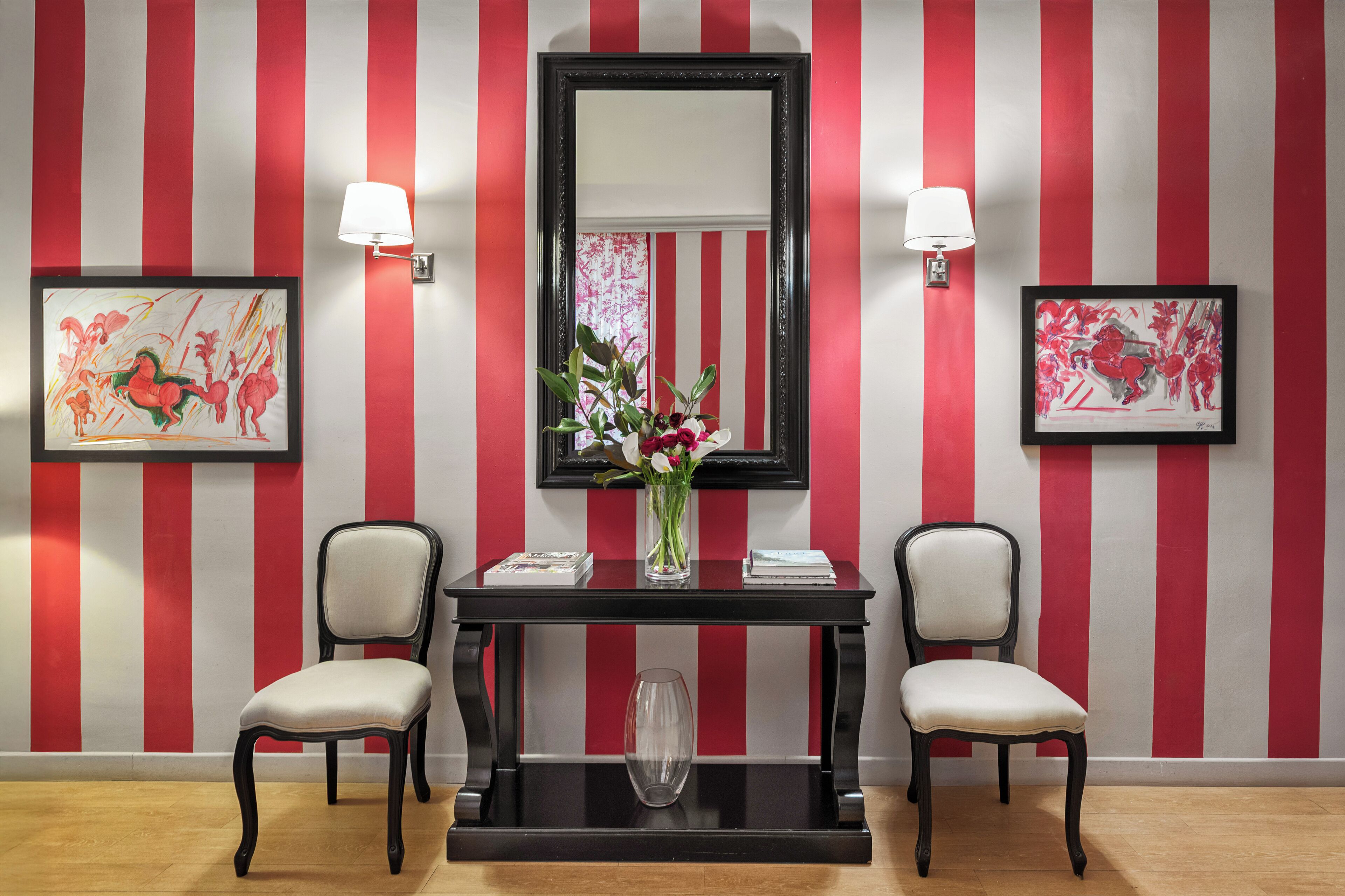 Une pièce avec un mur rayé de rouge et de blanc, un miroir à cadre noir, deux chaises, une petite table sur laquelle est posé un vase de fleurs et deux lampes fixées au mur.
