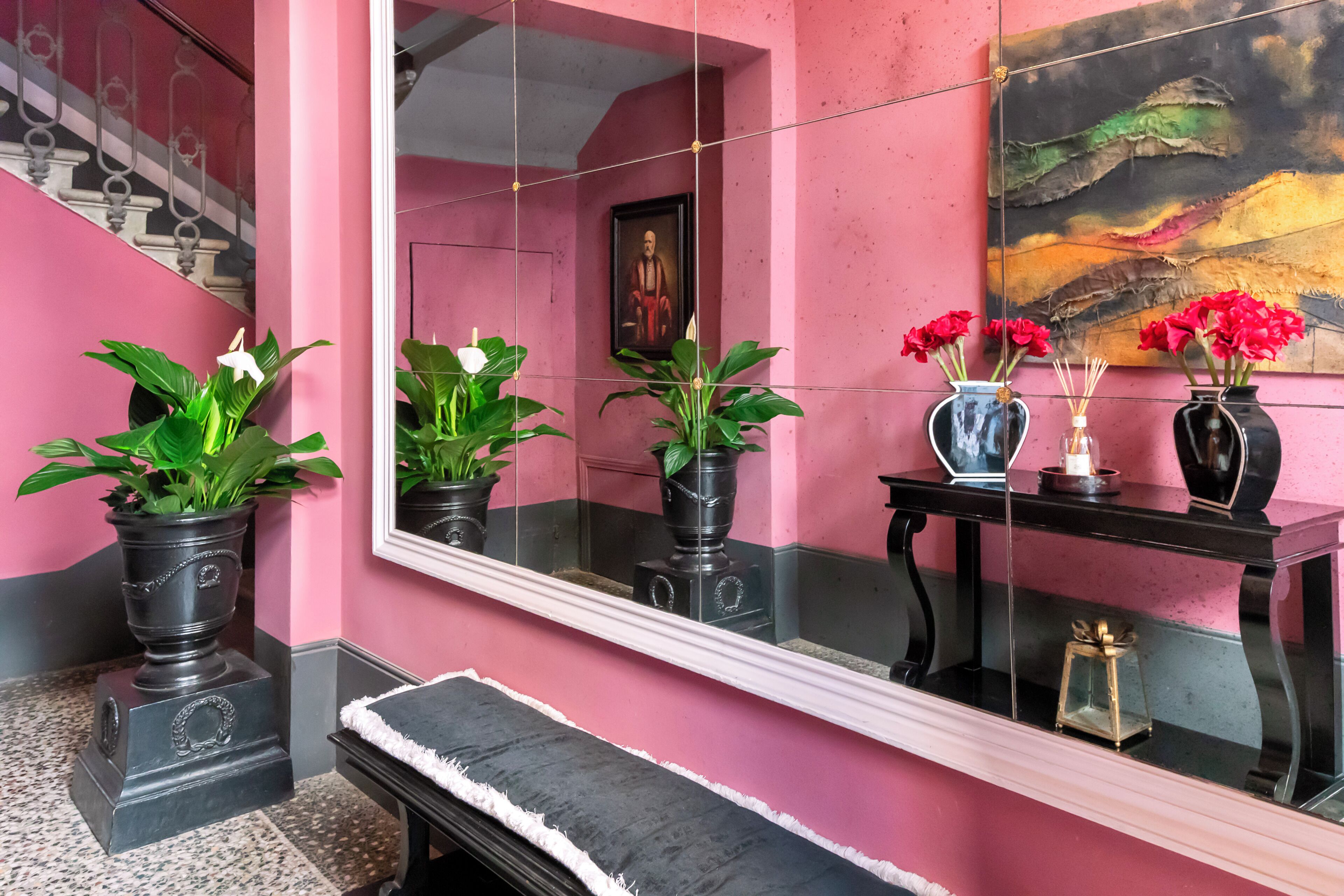 A pink-walled hallway with a large mirror reflecting a staircase and a painting. There are potted plants and a small table with vases and candles.