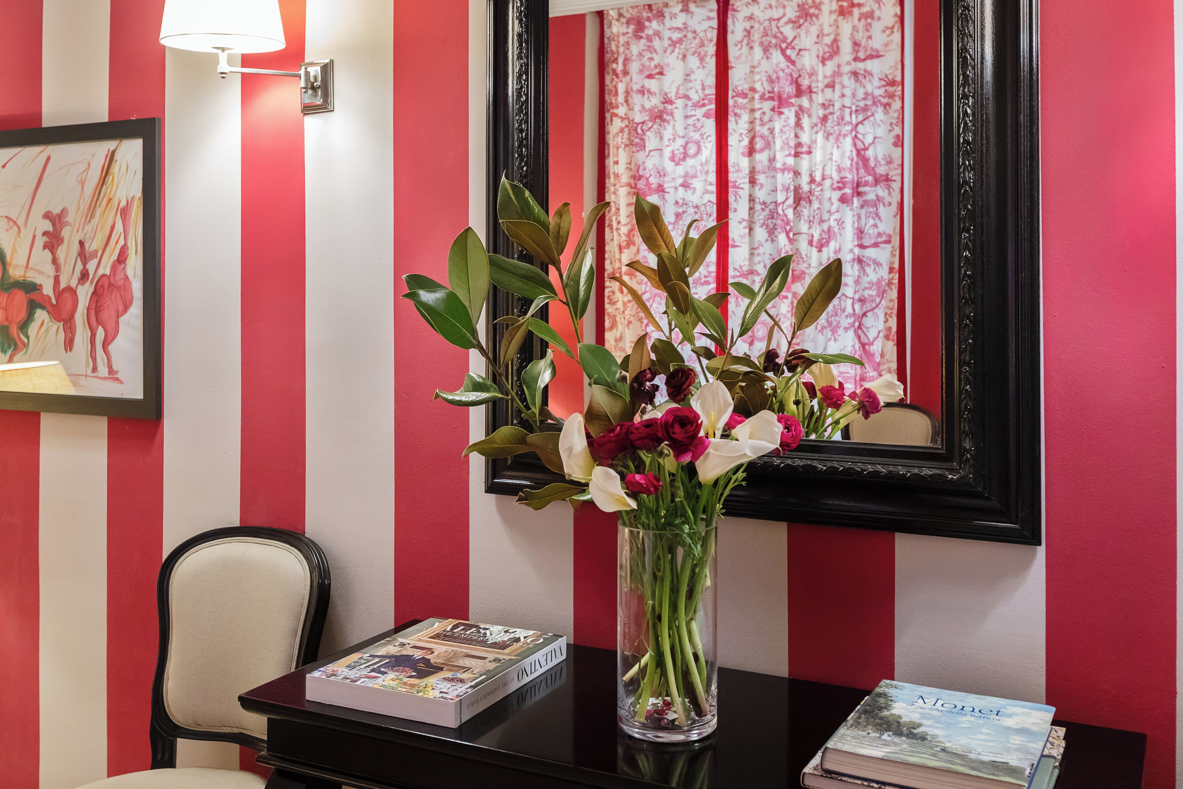 Une pièce avec un mur rayé de rouge et de blanc, un miroir encadré, un vase avec des fleurs et un livre posé sur une table.