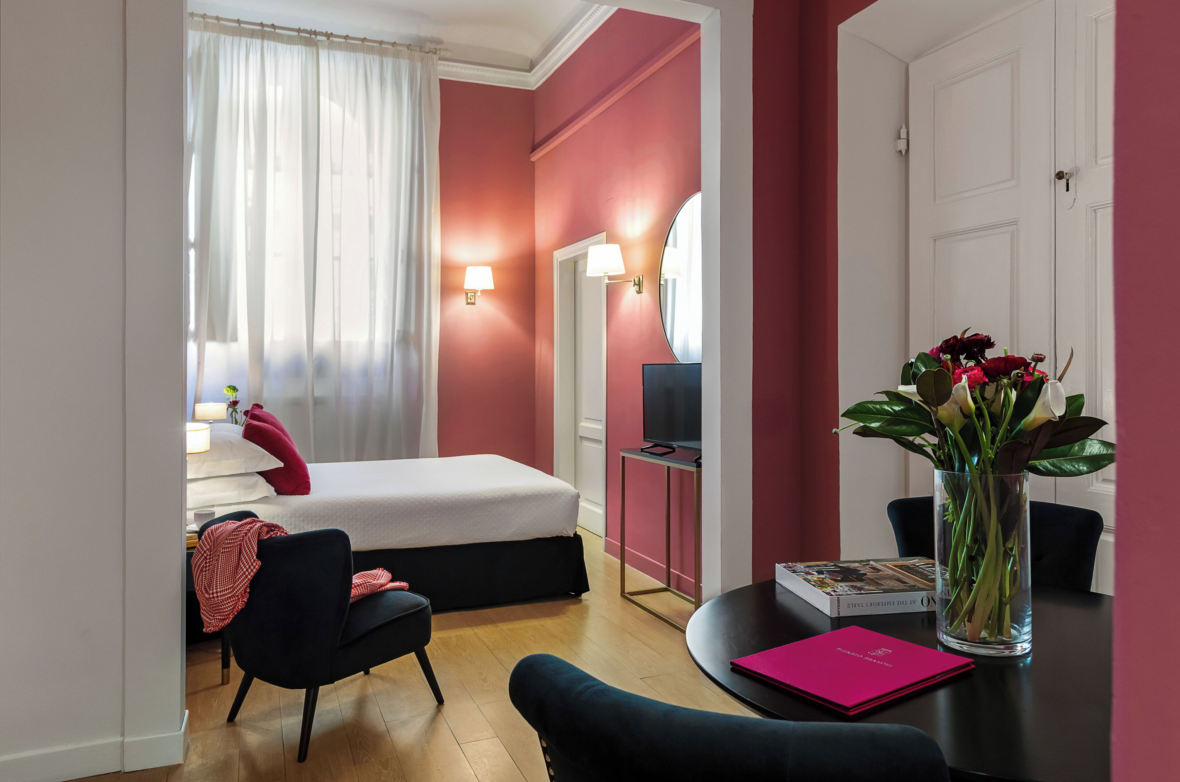 Ein Hotelzimmer mit einem Bett, einem Schreibtisch mit Computer, einem Stuhl, einem Tisch mit Blumenvase und einer Tür.