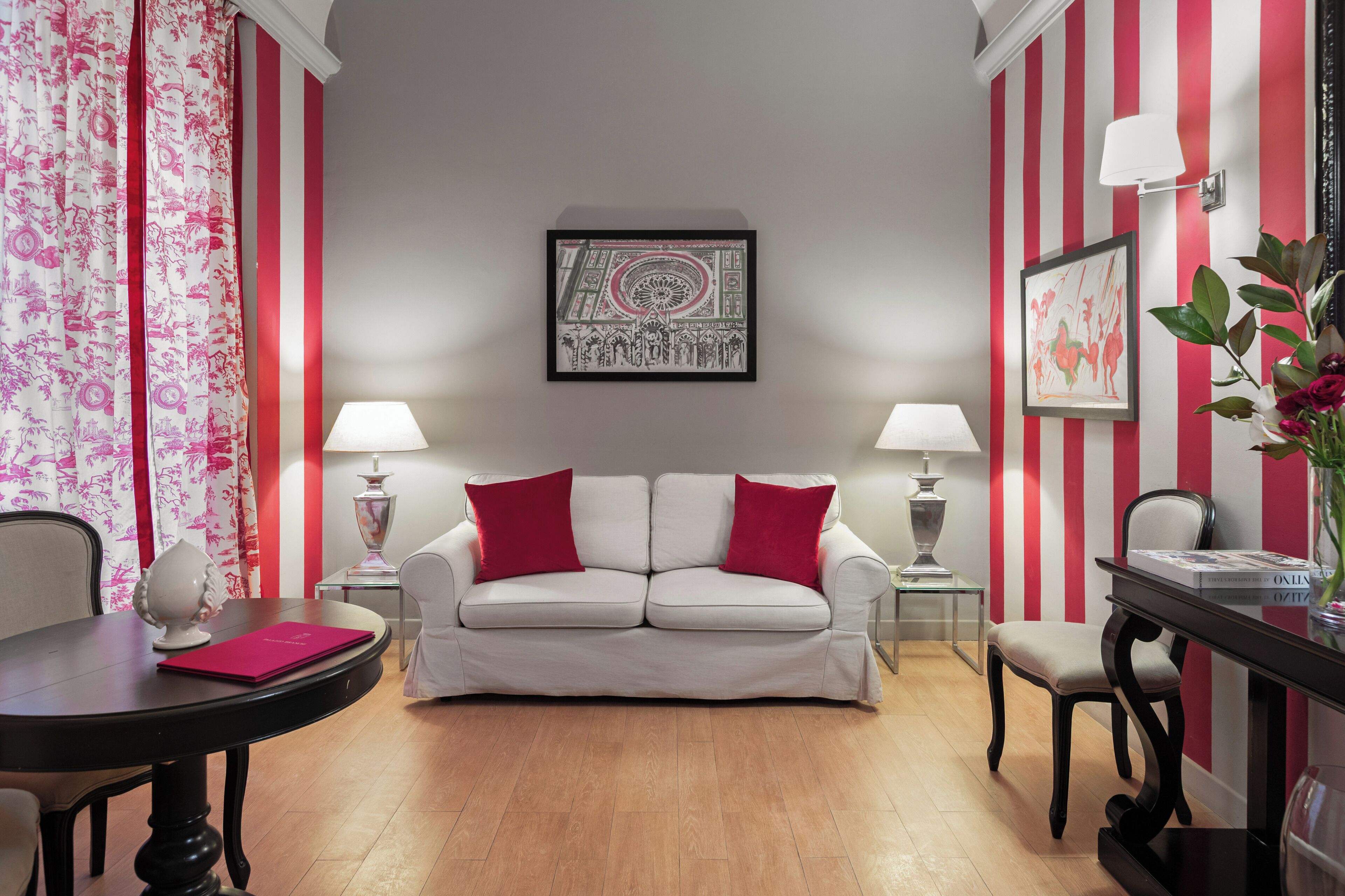 A room with a white sofa, two red cushions, a framed artwork on the wall, a table with a vase of flowers, and a striped wall.