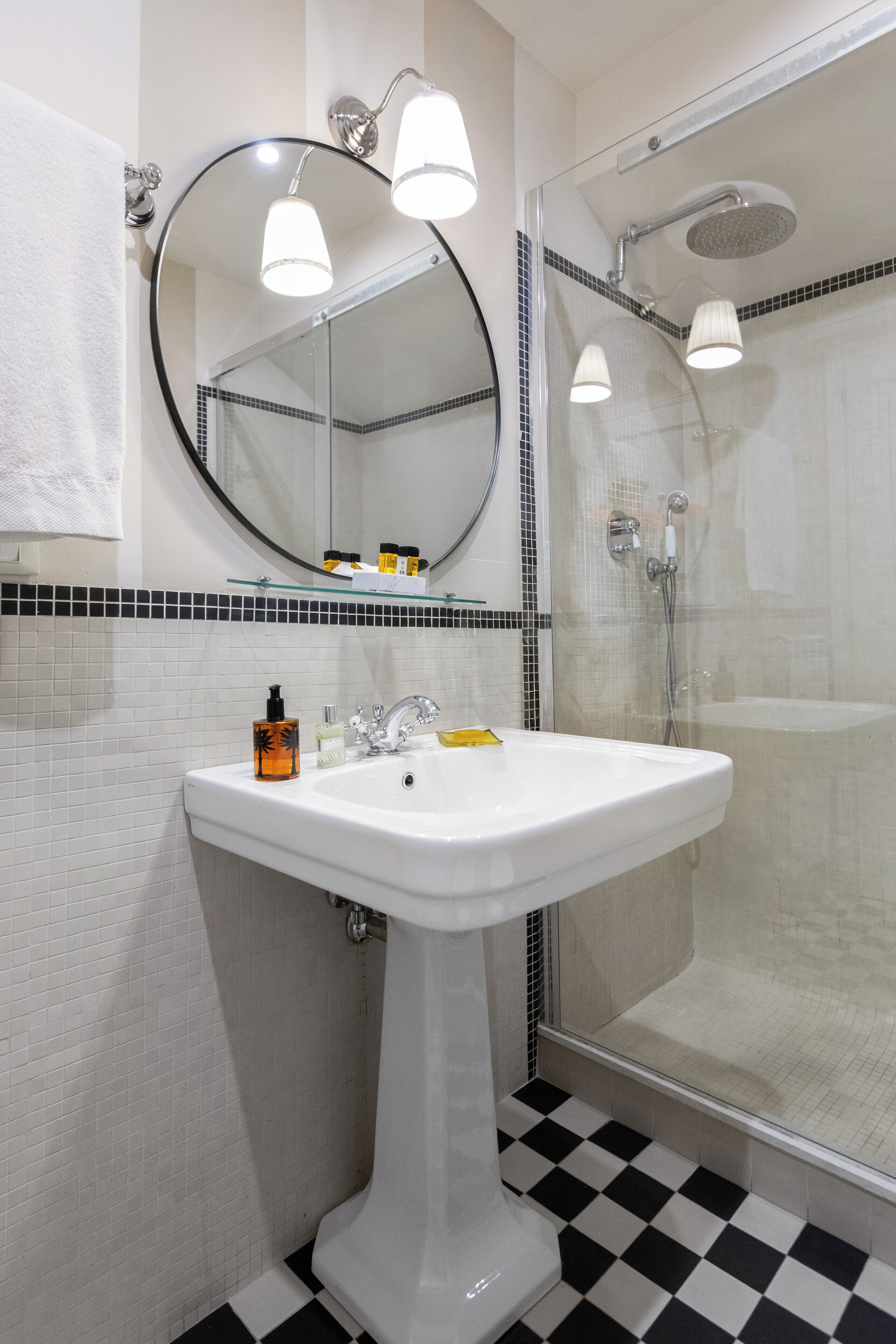 Une salle de bain avec un lavabo sur colonne blanc, un miroir rond et une douche séparée par une paroi de verre.