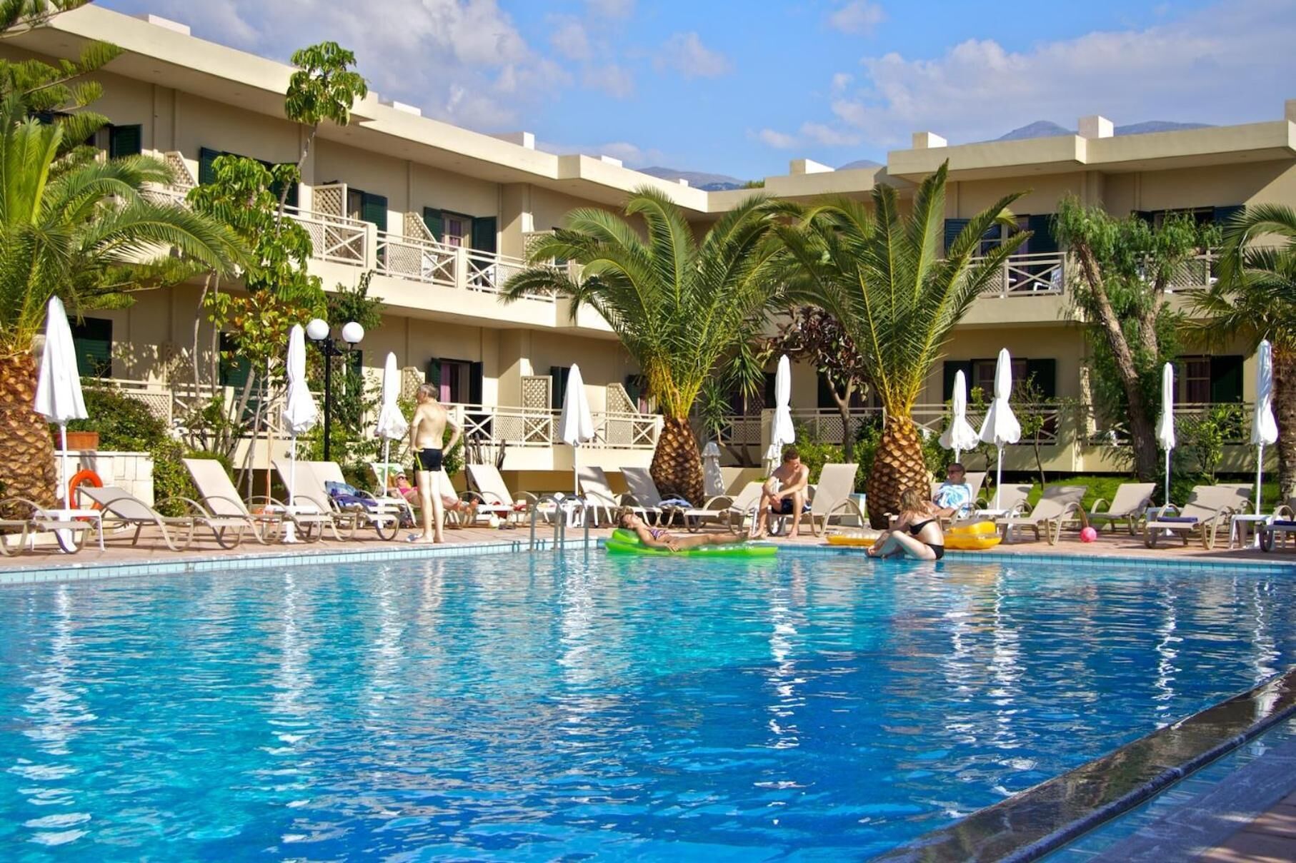 A pool area with lounge chairs and umbrellas, surrounded by palm trees and a multi-story building with balconies.