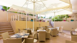 An outdoor dining area with wicker furniture, white umbrellas, and a staircase leading to a balcony with potted plants. - Solimar Ruby (Hersonissos)