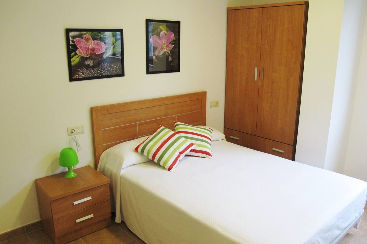 A neatly made bed with striped pillows, a wooden nightstand with a green lamp, and two framed pictures of flowers on the wall.