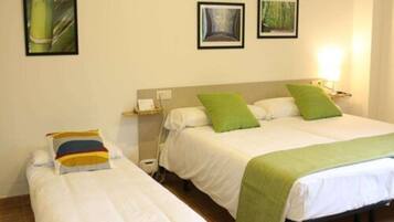 Habitación de hotel con dos camas, cada una con una almohada y una manta verde. Hay cuadros en la pared y una mesita de noche con una lámpara.