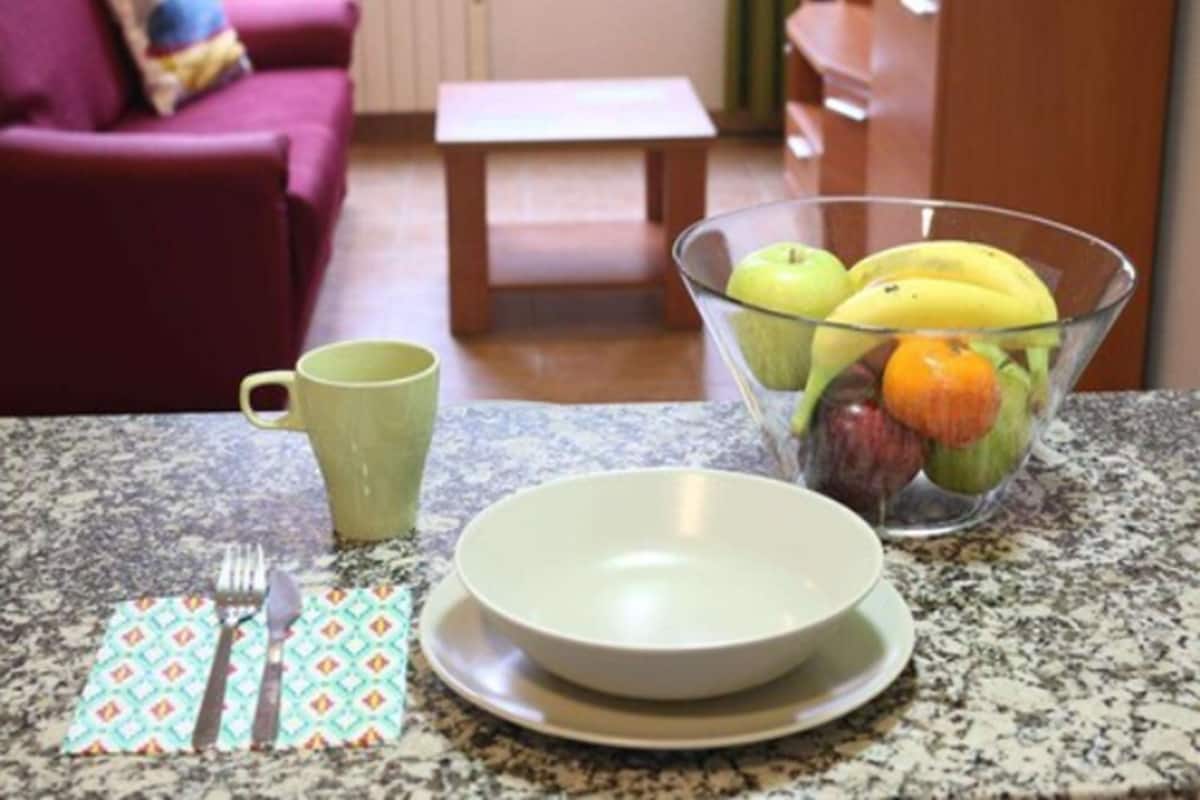 A kitchen counter with a bowl of fruit, a green mug, and a set of cutlery.