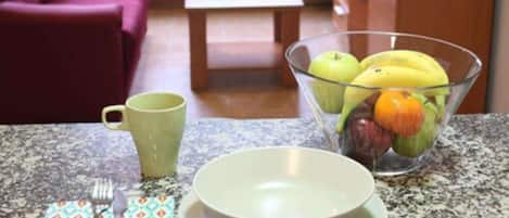 Una encimera de cocina con un bol de fruta, una taza verde y cubiertos.