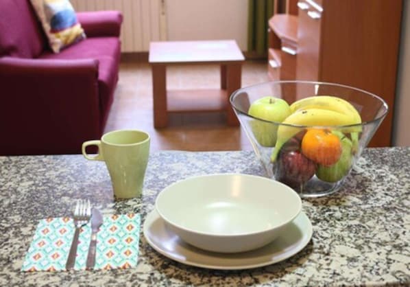 Una encimera de cocina con un bol de fruta, una taza verde y cubiertos.