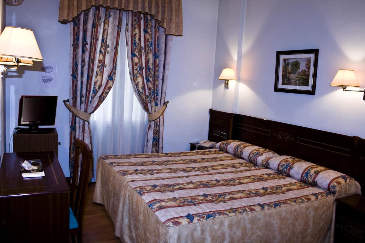 A hotel room with a bed, two bedside lamps, a desk with a lamp, a TV, and a chair.