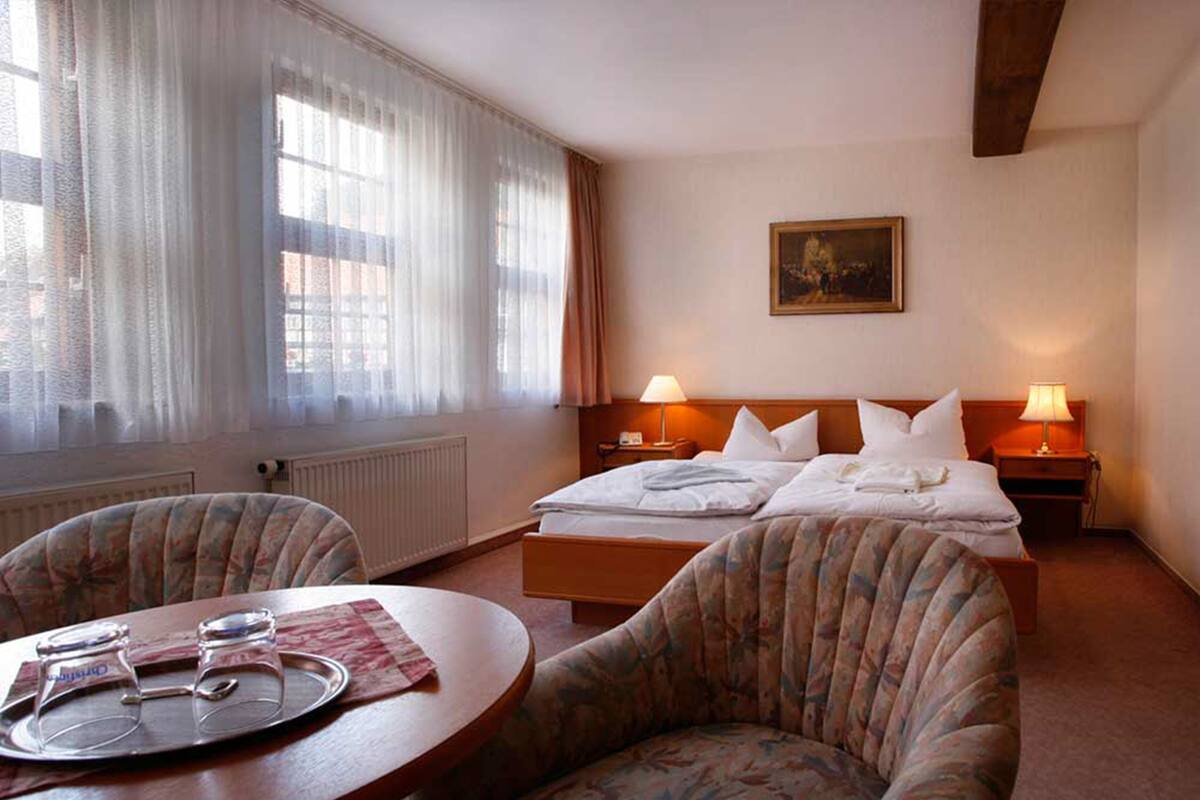 A hotel room with a bed, two bedside lamps, a painting on the wall, a table with a tray and glasses, and a chair.