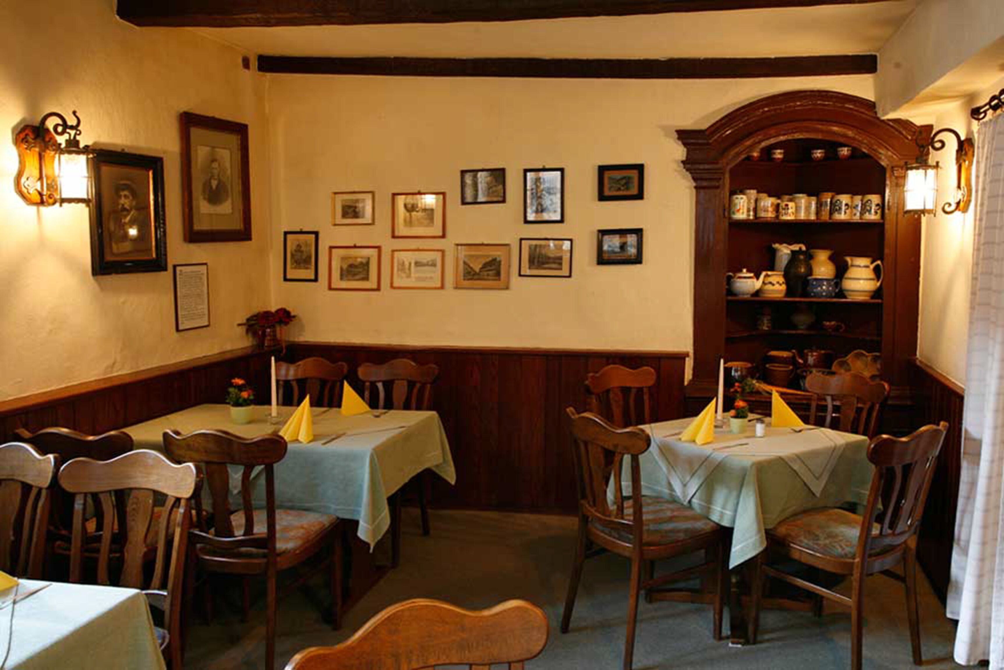 A dining area with wooden tables and chairs, framed pictures on the wall, and a shelf with kitchenware.