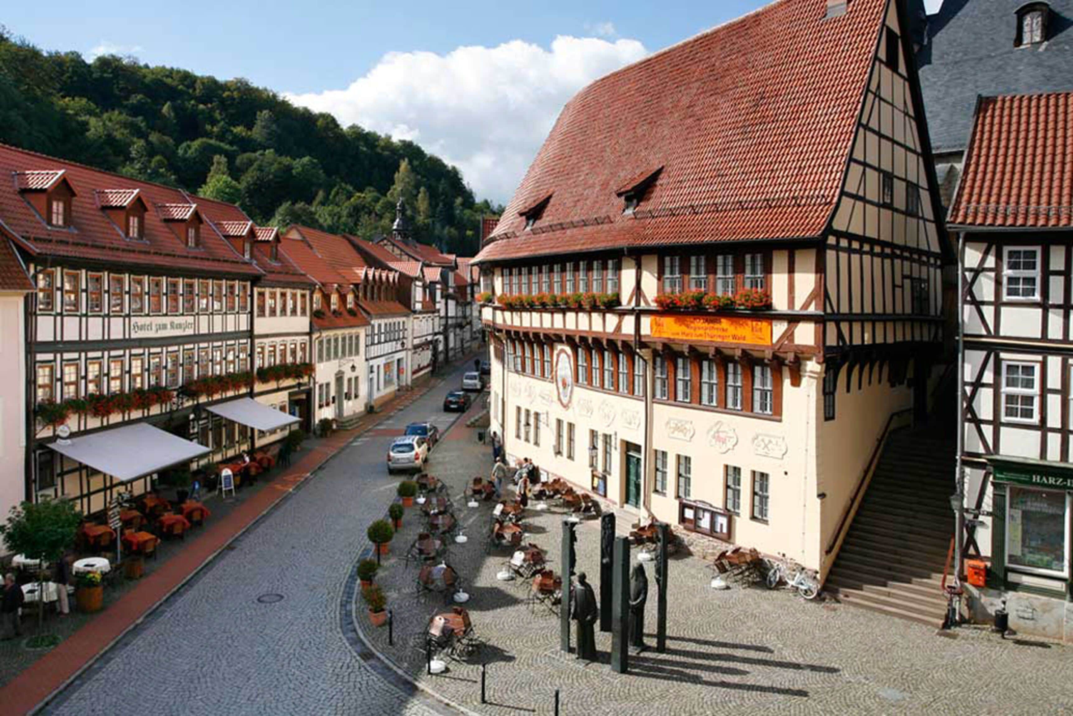 A traditional European town square with cobblestone streets, half-timbered buildings, outdoor seating for cafes, and a statue in the center.