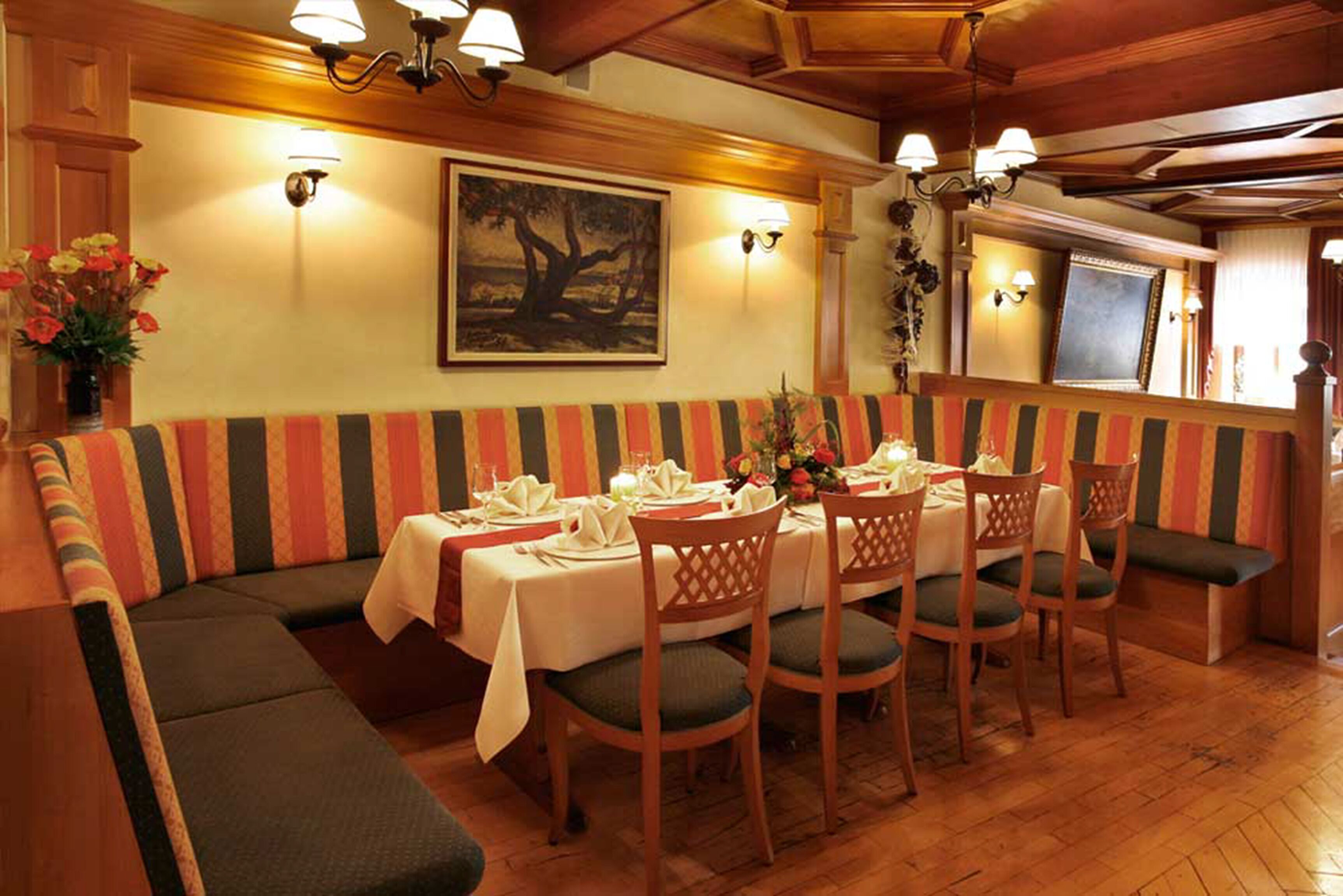 A dining area with a long table set for six, surrounded by wooden chairs with cushioned seats. A striped bench runs along the wall, and there are wall-mounted light fixtures above the table.