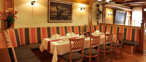 Un comedor con una mesa larga dispuesta para seis personas, rodeada de sillas de madera con asientos acolchados. Un banco a rayas recorre la pared, y hay lámparas fijadas a la pared sobre la mesa.