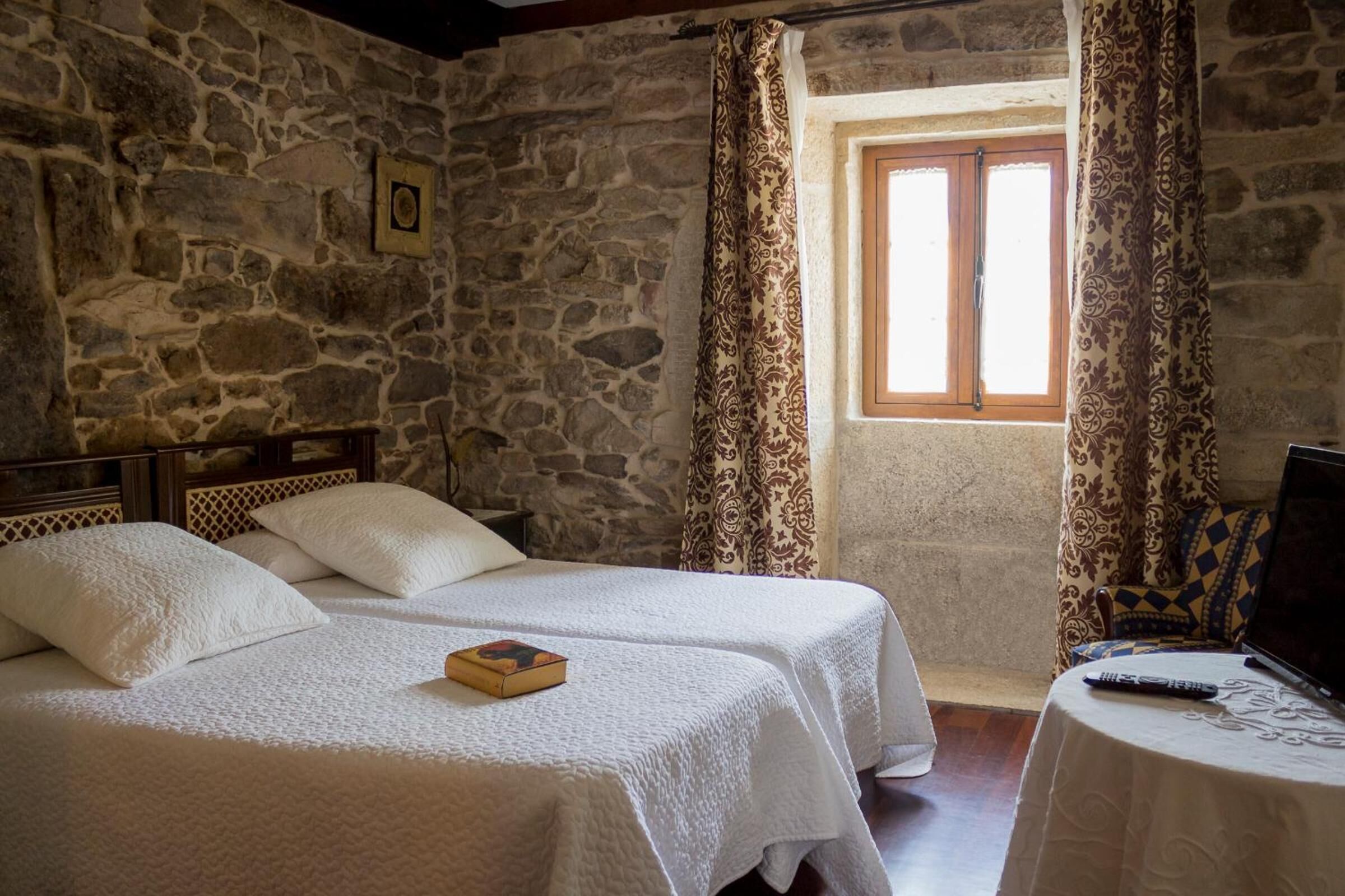 Una habitación con dos camas, paredes de piedra, un ventanal con cortinas y una mesita con un control remoto.