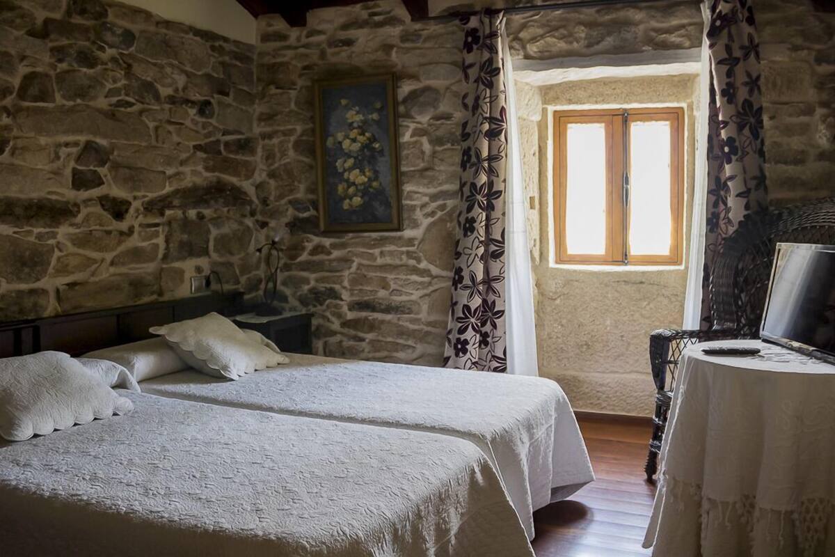 A room with two beds, a stone wall, a painting, a window with curtains, and a table with a TV.
