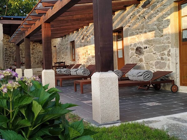 Un edificio de piedra con vigas de madera, mobiliario exterior con cojines y un jardín.