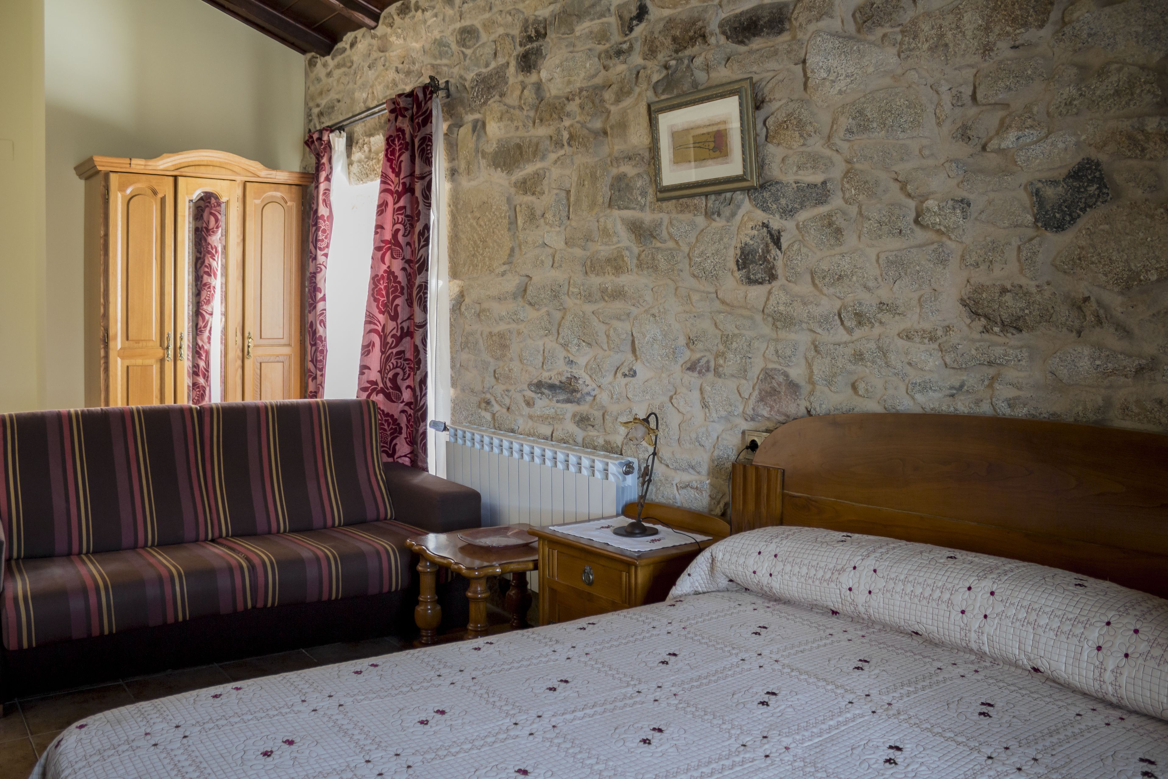 A room with a stone wall, a wooden bed, a striped sofa, a wooden cabinet, and a radiator.