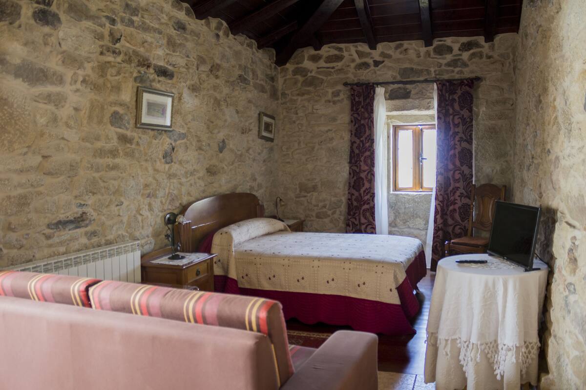 A stone-walled bedroom with a bed, a nightstand, a chair, a table with a laptop, and a window with curtains.