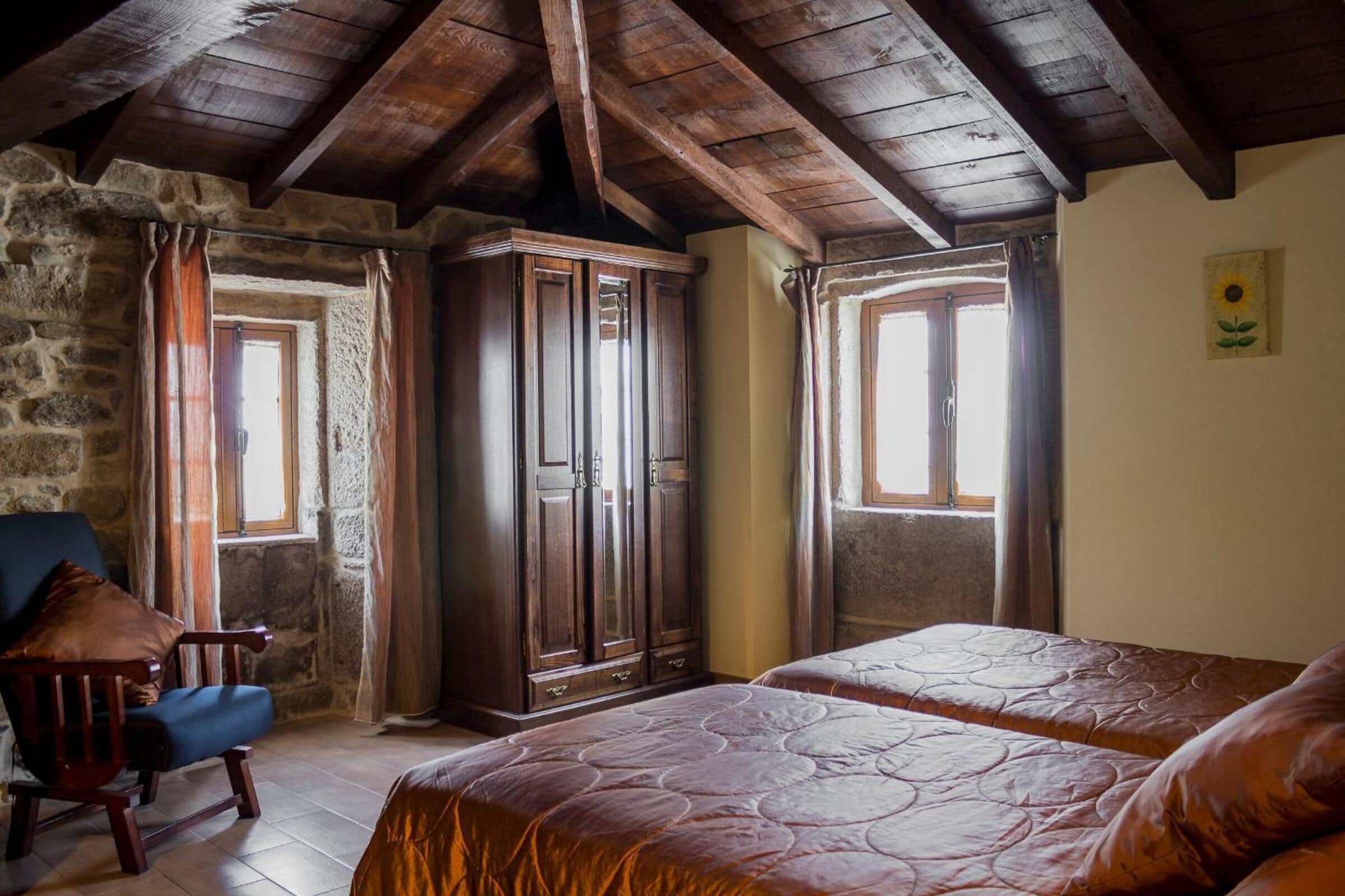 A rustic room with wooden beams, stone walls, two beds, a wooden wardrobe, and a chair.