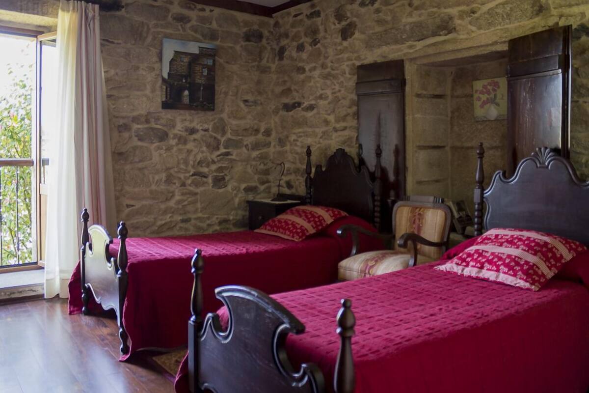 A room with stone walls, two wooden beds with red bedding, a wooden chair, and a window with curtains.