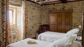 Un dormitorio con pared de piedra, armario de madera, dos camas con ropa de cama blanca, una araña y una silla.