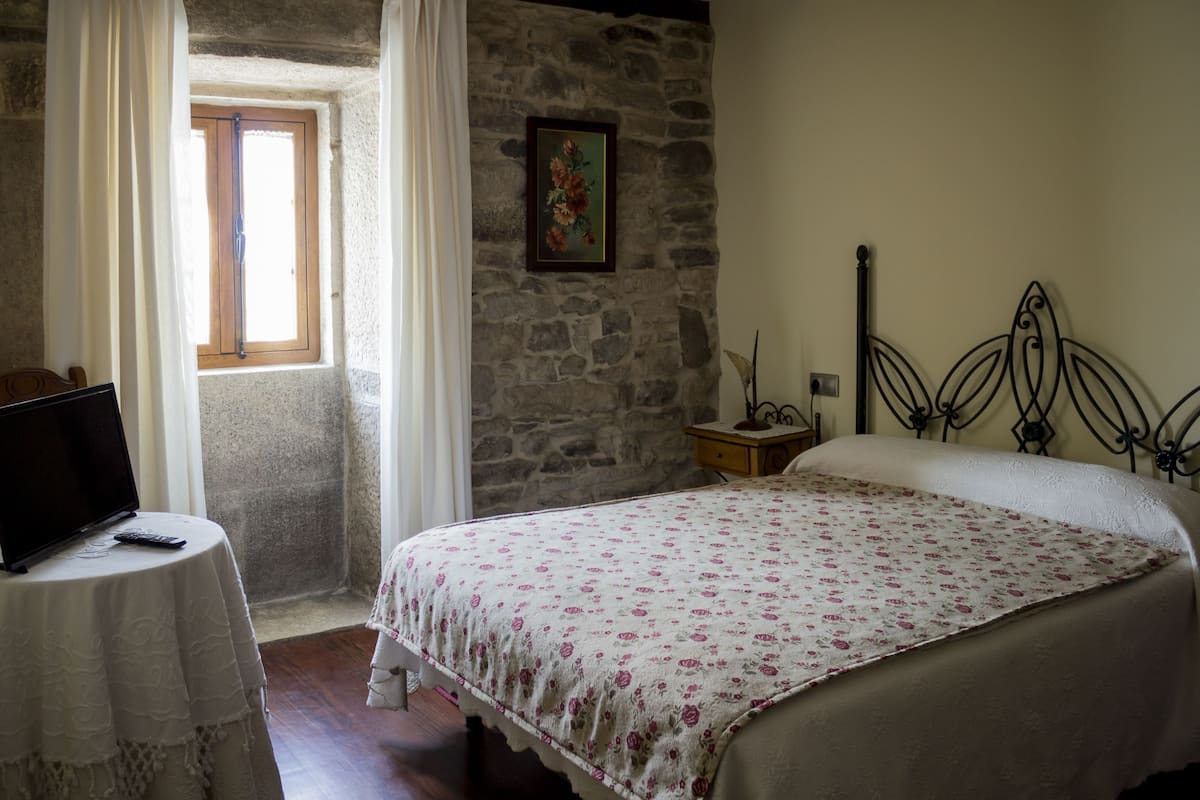 A bedroom with a stone wall, a bed with a floral bedspread, a bedside table, a lamp, and a window with curtains.