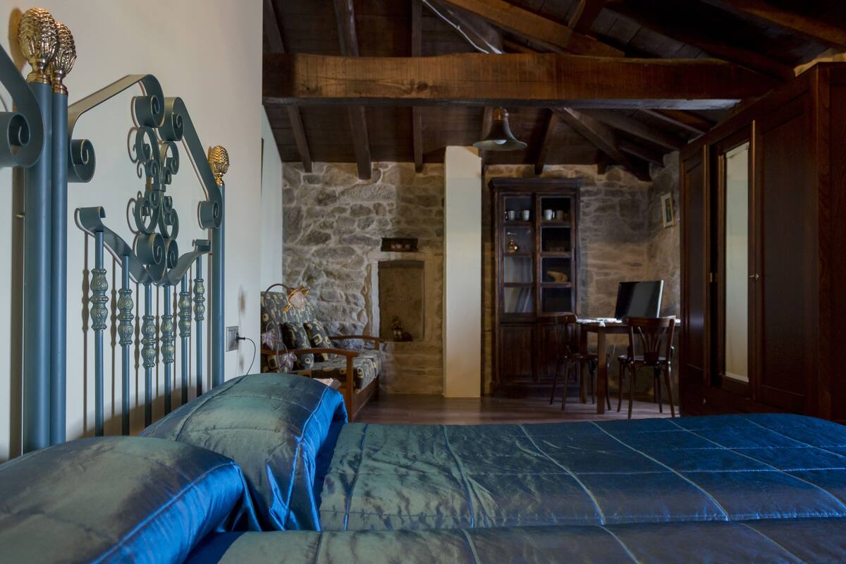 A bedroom with a bed, a wooden ceiling, a stone wall, and a desk with chairs.